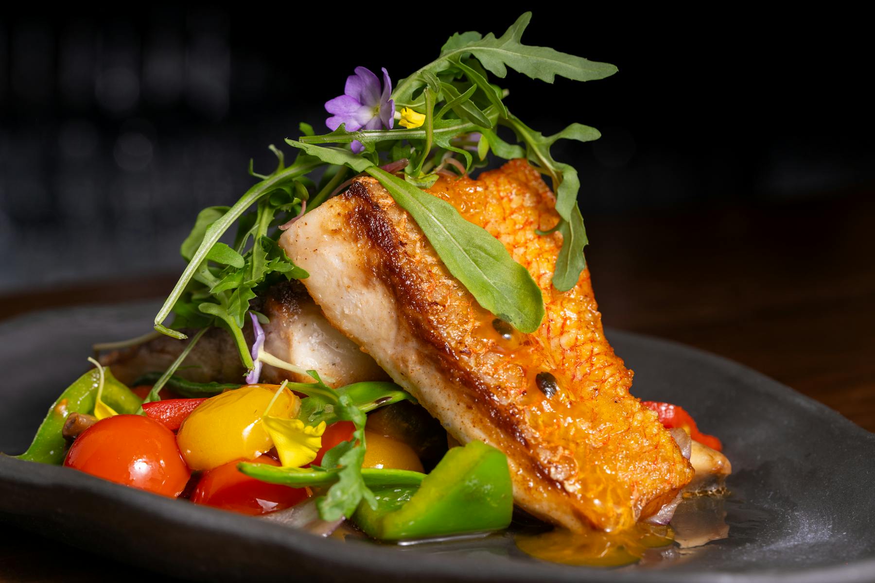 Delicious grilled fish topped with fresh vegetables and arugula on a dark plate.